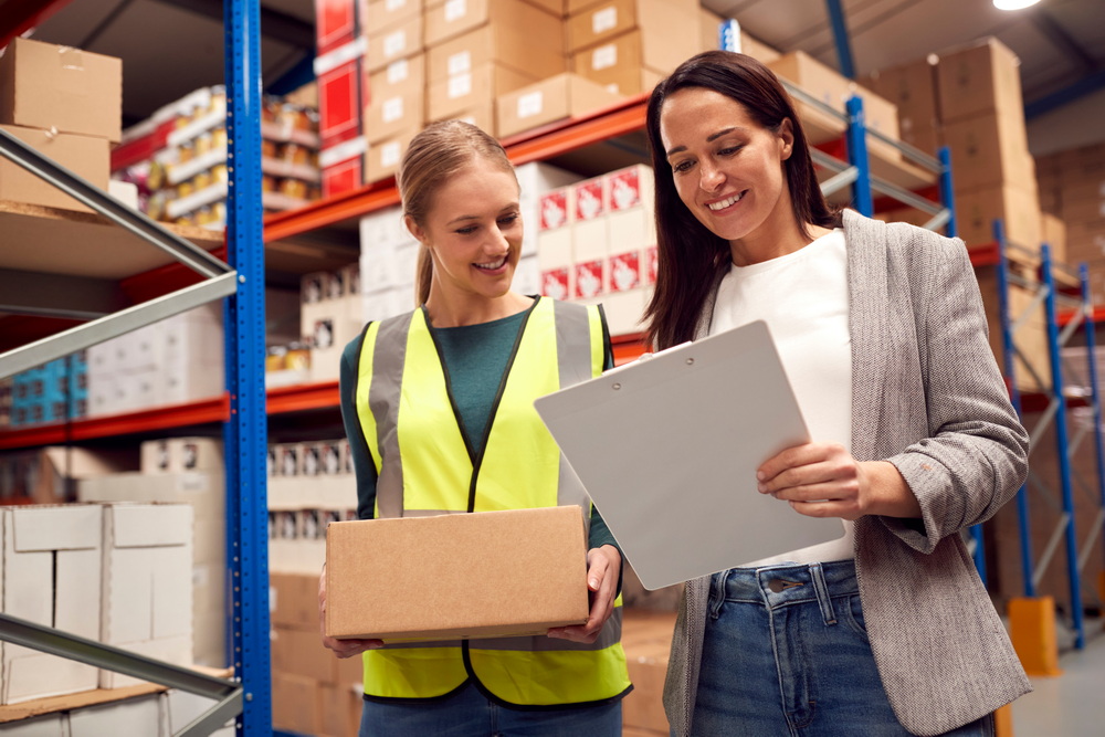 Female Team Leader With Clipboard In Warehouse Training Intern Standing By Shelves